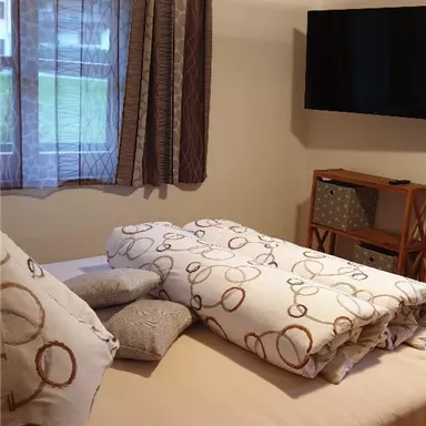 A cozy bedroom with a bed, pillows, and a blanket. A television hangs on the wall, and next to the bed is a small table.
