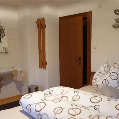 A cozy bedroom with a double bed and decorative cushions. There is a sink area and a shelf with towels.