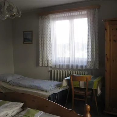 A simple room with two beds, a table, and a chair. The window lets in plenty of light and the walls are painted bright.