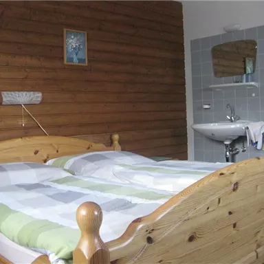 A simple bedroom with a wooden bed and a bright ceiling. In the background, there is a sink and a mirror.