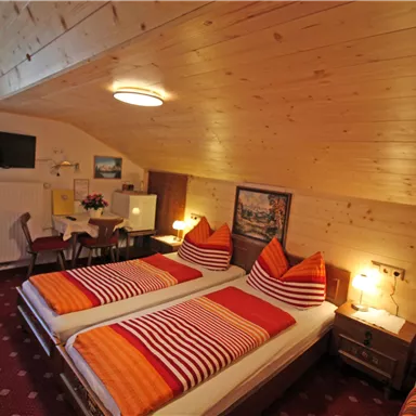 A cozy bedroom in the attic with two single beds and colorful pillows. The room is equipped with wooden walls, a television, and a small table.