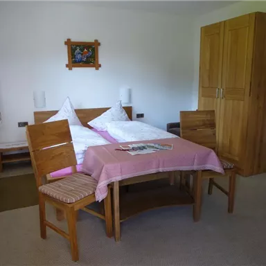 A simple bedroom with a bed, a table, and chairs. The room has a neutral tone and a window with curtains.