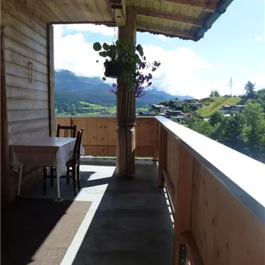 A balcony with a wooden railing and a table-chair combination. In the background, hills and mountains are visible beneath a clear sky.