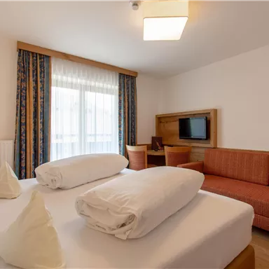 A modern room with a double bed, two pillows, and a cozy sofa. The room has large windows that let in plenty of daylight.
