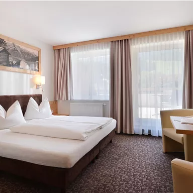 Ein modernes Hotelzimmer mit einem Doppelbett und zwei großen Fenstern. Der Raum ist gemütlich mit einem Tisch und einem Fernseher ausgestattet.