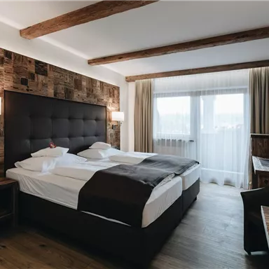 A modern hotel room with a large double bed and wooden elements. Bright windows create a friendly atmosphere.