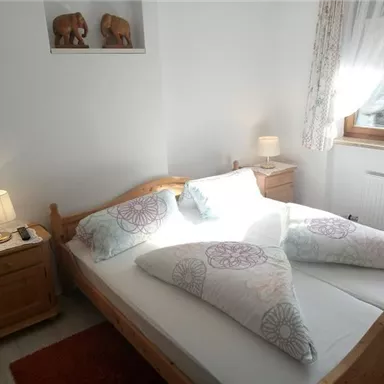 A cozy bedroom with a large bed and decorative pillows. Wooden figures can be seen on the walls, and the light from a lamp creates an inviting atmosphere.