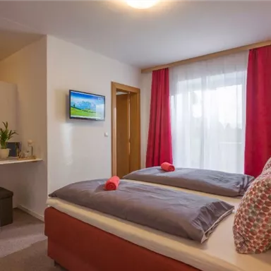 A cozy bedroom with two beds, a TV, and a small table. Large windows let in plenty of light, and red curtains add warmth to the room.