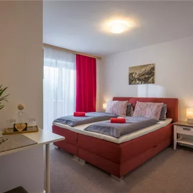 A cozy bedroom with two beds, red accents, and a large window front. The room is well-lit and modernly furnished.