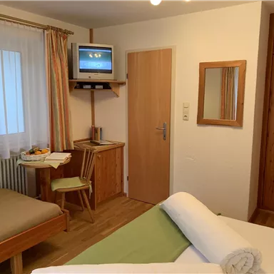 A cozy hotel room with a bed, wooden shelves, and a television. In the room, there is a table with chairs and a window with curtains.
