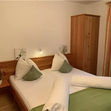 A cozy bedroom with two beds, pillows, and a wooden dresser. The room has bright walls and decorations in soft colors.