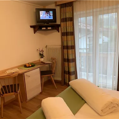 A cozy hotel room with a table, chairs, and a television on the wall. Large windows provide plenty of daylight.