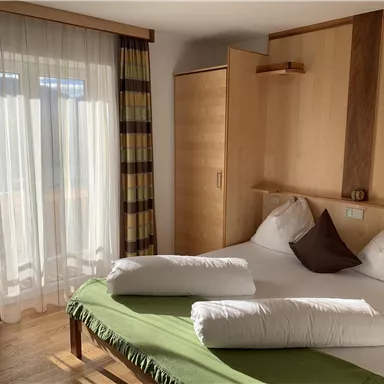 A cozy bedroom with a large bed and bright, natural wood colors. Light curtains hang in front of a window, and a green blanket is laid on the bed.