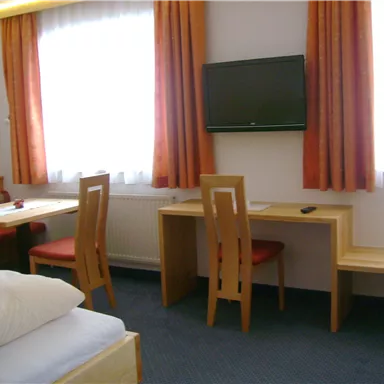 A simple room with a bed, a table, and chairs. There is a television and bright windows with curtains.