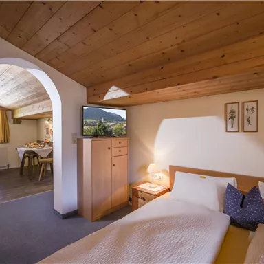 Ein gemütliches Schlafzimmer mit Holzdecke und zwei Betten. Im Hintergrund sieht man einen Wohnbereich mit Esstisch und Fernseher.