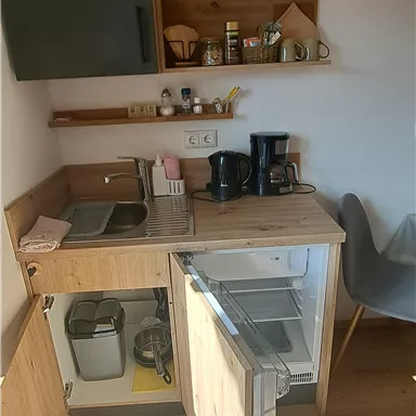 A small kitchenette with wooden shelves and a coffee maker. The refrigerator is open and there is a bright, friendly atmosphere.