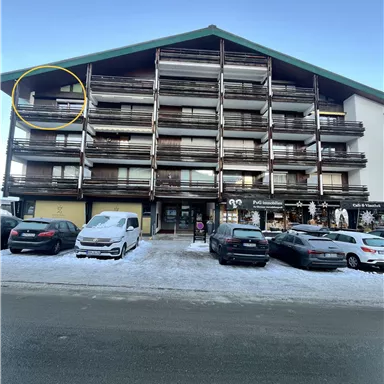 A modern residential building with several floors and balconies. In front of the building, cars are parked on a snow-covered street.