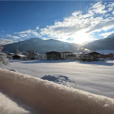 A winter landscape with snow-covered fields and mountains. The sun shines through the clouds and illuminates the scene.