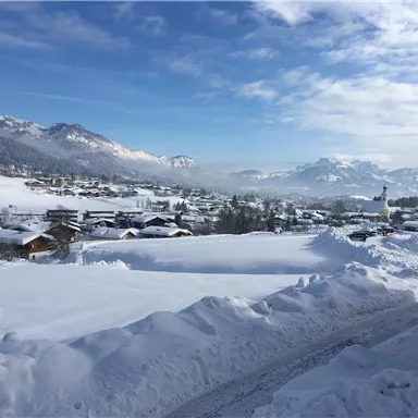 A picturesque winter landscape with snow-covered hills and small houses. The sky is clear and the mountains are visible in the distance.