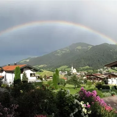A breathtaking view of the mountains with a shining rainbow. In the foreground, colorful flowers are blooming and there are charming houses.