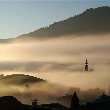 A misty landscape with gentle hills and a church tower in the background. The warm morning atmosphere invites reflection.