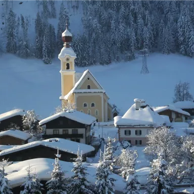 Eine malerische winterliche Landschaft mit verschneiten Hütten und einer Kirche. Die Bäume sind mit Schnee bedeckt und die Umgebung strahlt Ruhe aus.