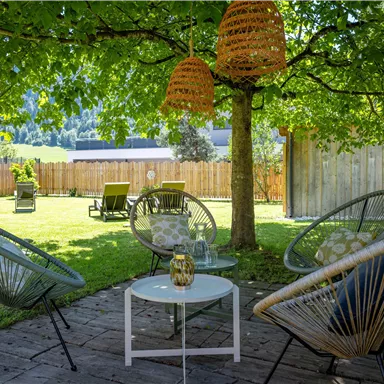 A cozy outdoor area with several chairs and a table under a tree. The surroundings are green and inviting, perfect for relaxing hours outside.