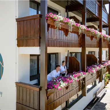 A modern hotel building with balconies adorned with colorful flowers. Two people are standing on a balcony, enjoying the view.