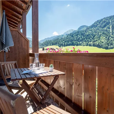 A cozy balcony with a small table and chairs. In the background, green mountains and a clear blue sky are visible.