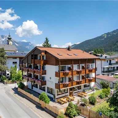 A charming hotel with wooden cladding and balconies, surrounded by green nature. In the background, majestic mountains and a blue sky can be seen.