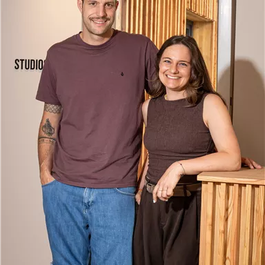 A man and a woman are smiling next to each other in a modern room. In the background, there is a wood-paneled wall that creates an inviting atmosphere.