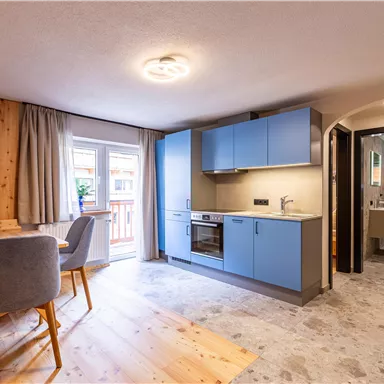 A modern kitchen with blue cabinets and a dining area. The space has a bright, friendly atmosphere with wooden details and plenty of daylight.