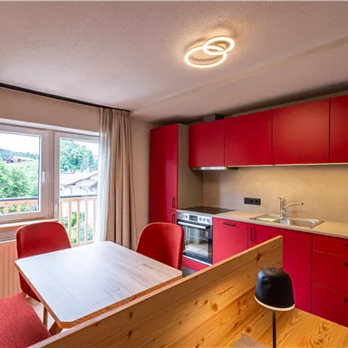 A modern kitchen with red cabinets and a small dining area. The window offers a view of the surroundings.