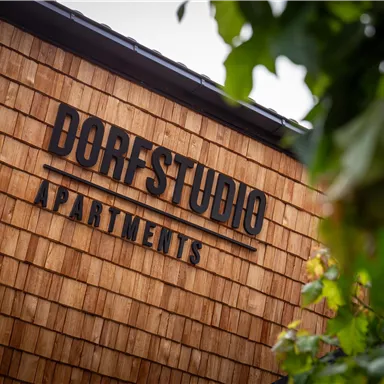 A modern building with a wooden facade and a corner surrounded by plants. The inscription "Dorfstudio Apartments" is clearly visible.
