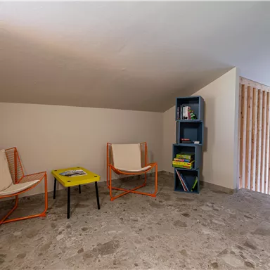 A modern, bright room with two chairs and a colorful table.
On the wall stands a shelf with books, and light falls through a window.