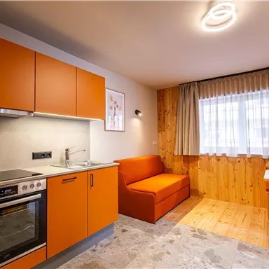 A modern kitchenette with orange cabinets and a cozy living area. The light from above and the window create a bright atmosphere.