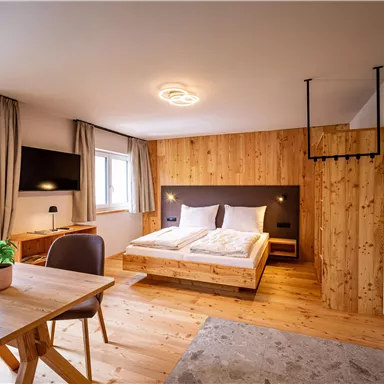 A modern room with wooden decor, a double bed, and a table. Large windows provide plenty of daylight and a cozy atmosphere.