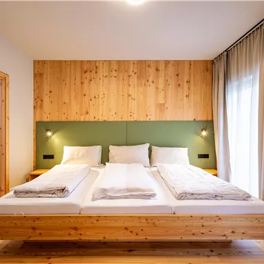 A modern bedroom with a large wooden bed and two pillows. The walls are painted in soft colors, and large windows let in plenty of light.