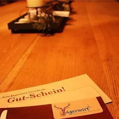 A wooden table with a gift voucher and a menu card. In the background, there is a candle on a decorated tray.