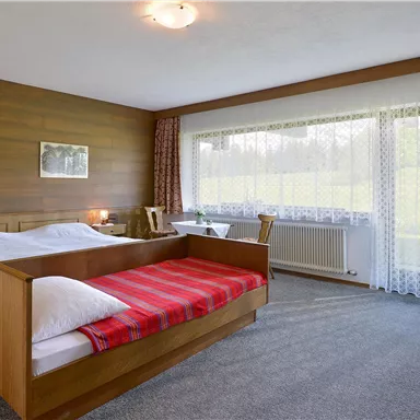 A simple bedroom with two beds and wooden furniture. Large windows provide plenty of daylight and views of the surroundings.