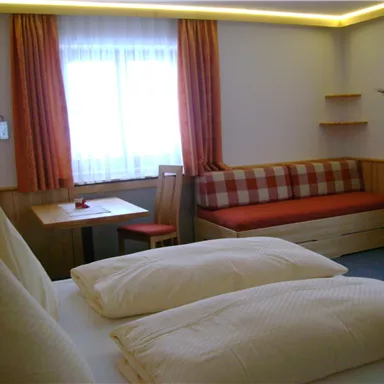A cozy guest room with two beds, a table, and a seating area. The room has windows with daylight and orange curtains.