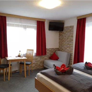 A cozy room with two beds, a sofa, and a table. The windows let in plenty of daylight and the red curtains give a warm atmosphere.