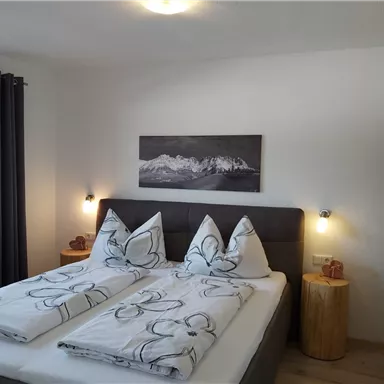 A cozy bedroom with a double bed and modern wooden furniture. The walls are light and decorated with a landscape painting.