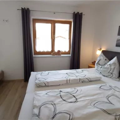 A cozy bedroom with two beds and bright windows. The walls are painted in neutral colors, and there is decorative bedding with a floral pattern.