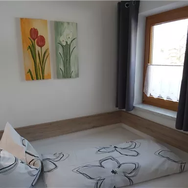 A cozy bedroom with a single bed and floral bedding. Colorful flower pictures hang on the wall, and a window lets in plenty of natural light.