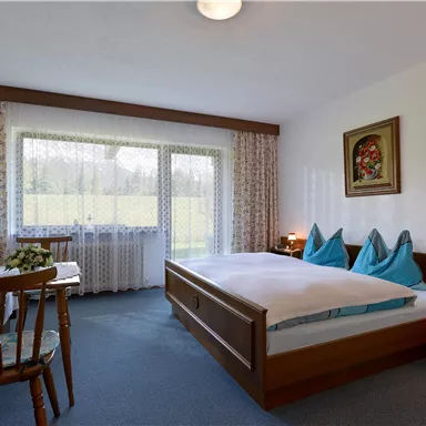 A cozy bedroom with two beds and blue pillows. Large windows offer a view of the green landscape.