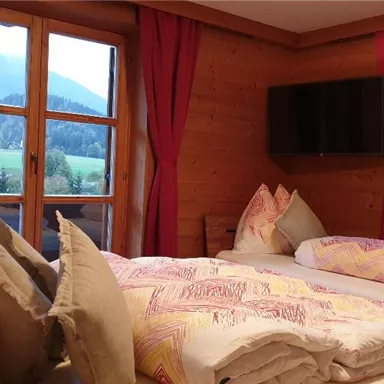 Cozy bedroom with wooden walls and large windows. The beds are decorated with colorful pillows and blankets, and the view shows a green landscape.
