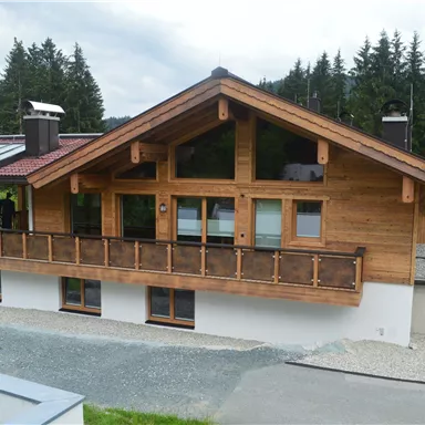 Ein modernes Holzhaus in den Bergen mit einem großen Balkon. Im Hintergrund sind Nadelbäume und eine bewölkte Himmel zu sehen.