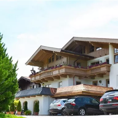 A charming alpine building with wooden decoration and balconies. Several cars are parked in front of the house, and there is plenty of green surroundings.