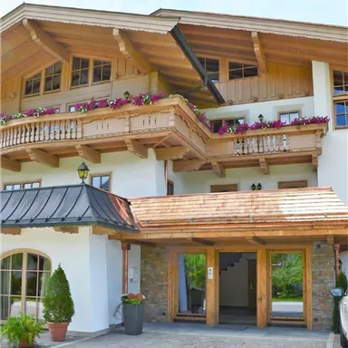 A cozy wooden house with balconies and colorful flowers. The front view shows the entrance area and a paved driveway.
